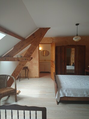 Un dormitorio con techo inclinado, vigas de madera, cama, sillón, cómoda y espejo.