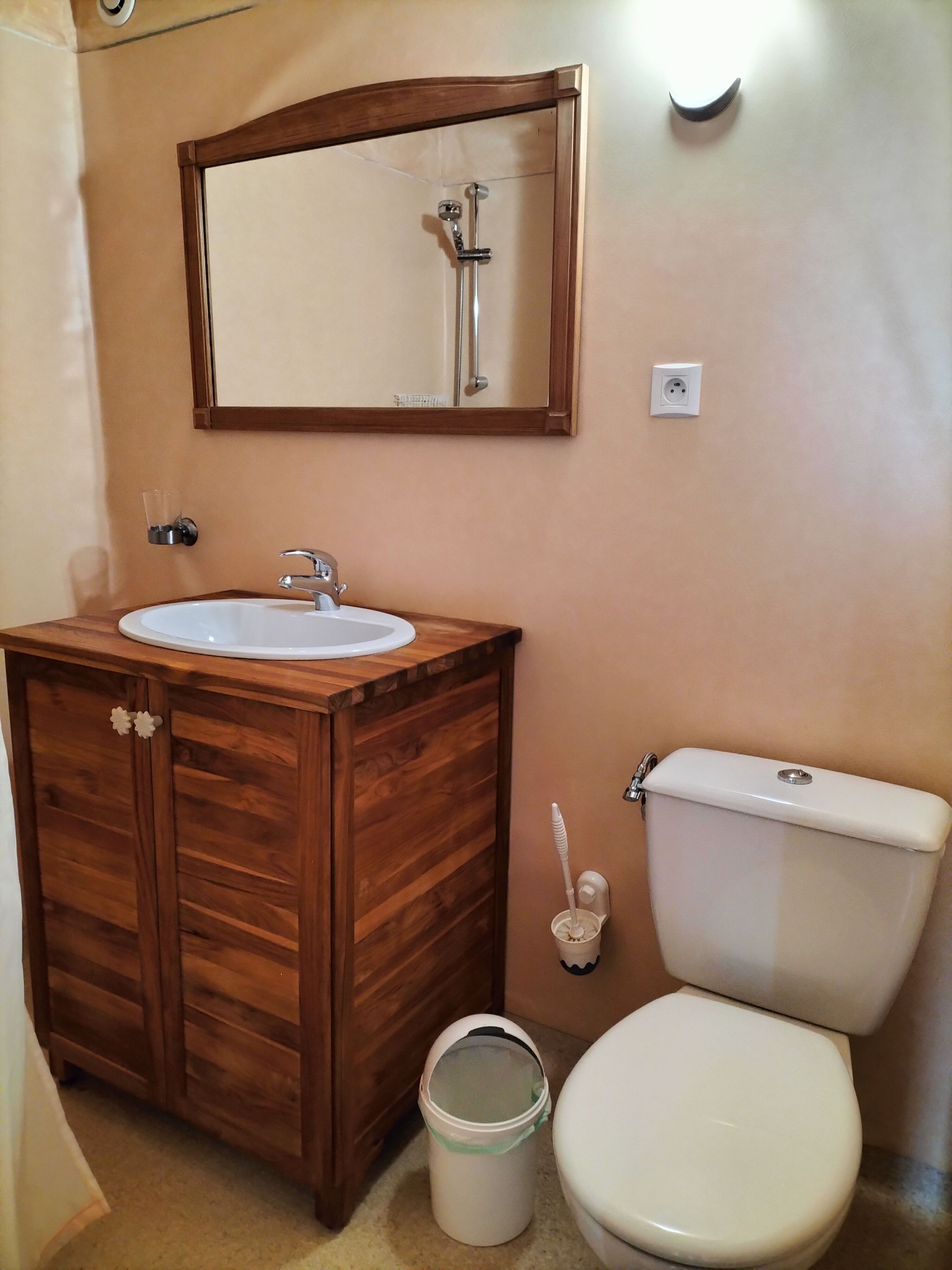 Een badkamer met een houten wastafel, een wasbak, een spiegel, een toilet en een emmer.