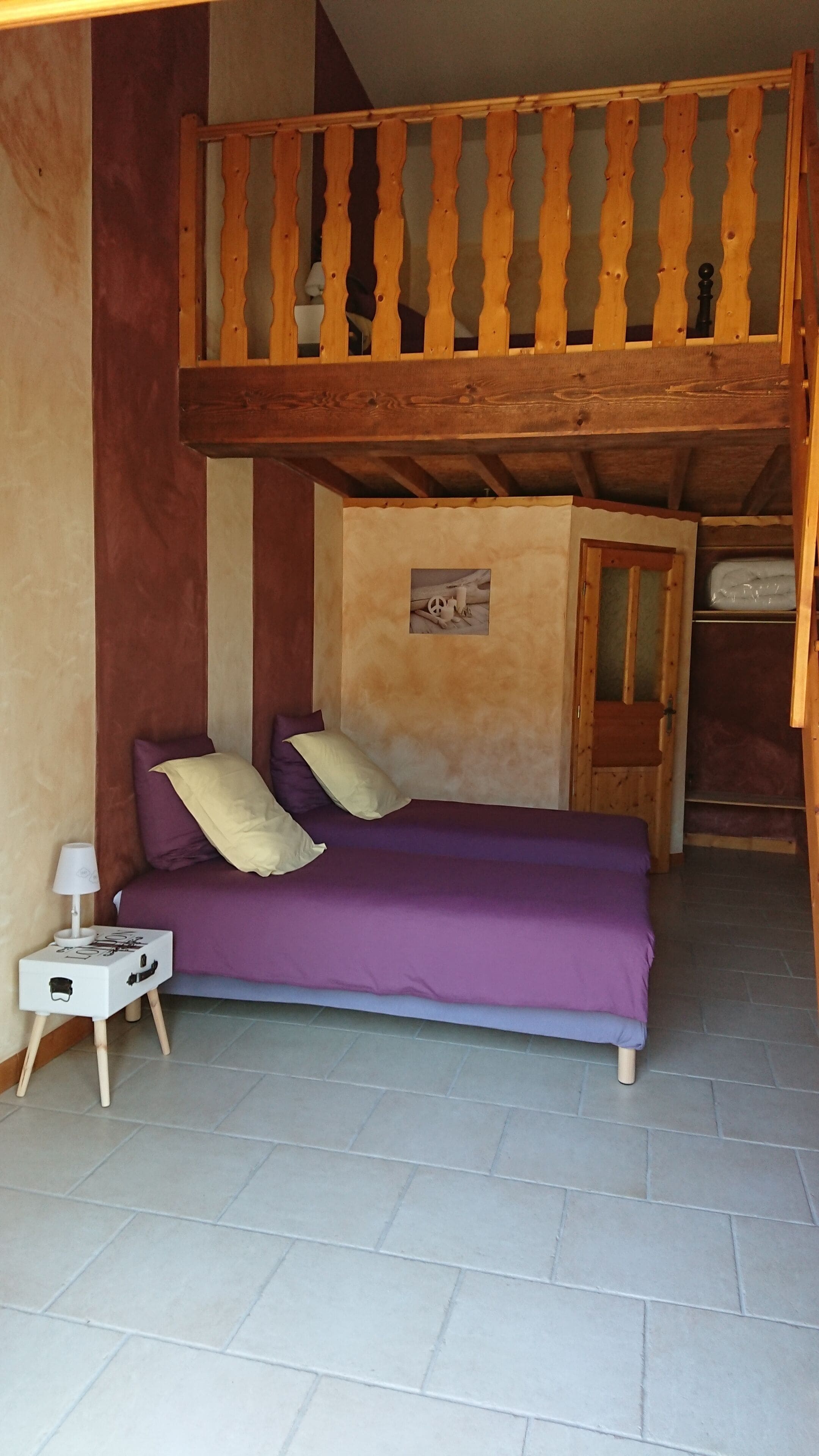 Ein Zimmer mit einem Etagenbett, einem Einzelbett mit violetter Bettwäsche, einem kleinen Nachttisch mit Lampe und einem an der Wand montierten Regal.