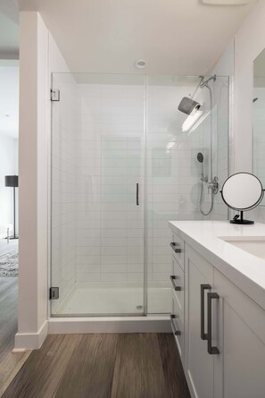 A modern bathroom with a glass-enclosed shower, a white vanity with a round mirror, and a wooden floor. - NEW 3br Luxury Townhome | Gaslamp | Balboa Park (San Diego)