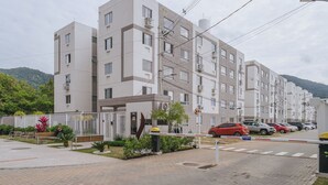 Un immeuble résidentiel à plusieurs étages, doté d’une entrée sécurisée par un portail, d’un espace de stationnement où sont garées plusieurs voitures, et d’un espace paysager agrémenté de bancs et de plantes.