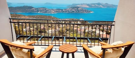 A balcony with two wooden chairs and a small table, overlooking a coastal landscape with hills and a body of water.