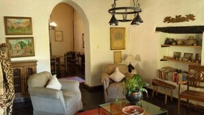 Ένα σαλόνι με έναν καναπέ, μια πολυθρόνα και ένα τραπεζάκι σαλονιού με ένα φυτό. Υπάρχει επίσης μια βιβλιοθήκη με βιβλία και διακοσμητικά αντικείμενα.