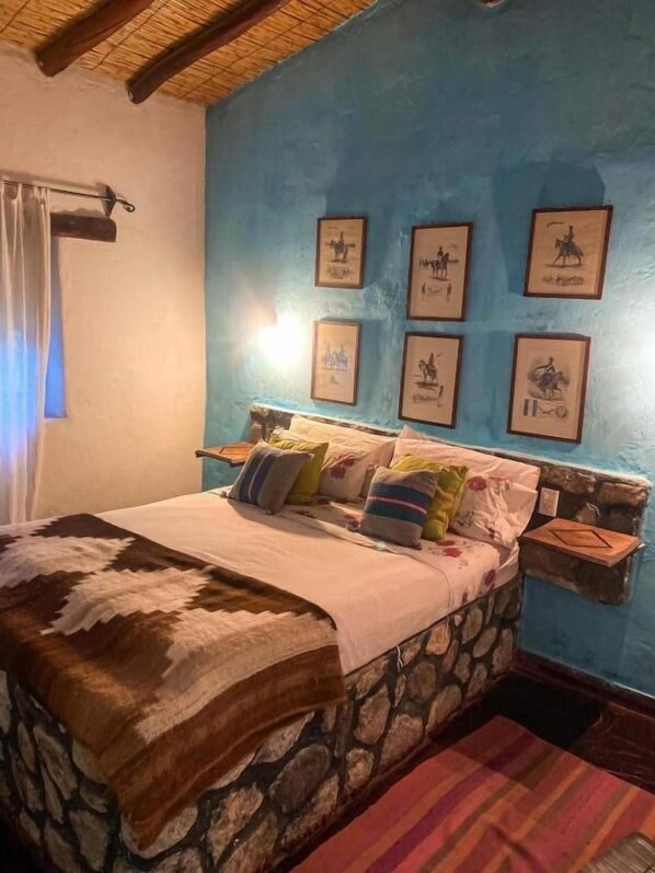Een bed met een hoofdbord van steen, een blauwe wand met ingelijste afbeeldingen en een houten plafond.