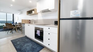 A modern kitchen with white cabinetry, a stainless steel refrigerator, and a built-in oven. There is a dining area with a table and chairs, and a view of the outdoors.
