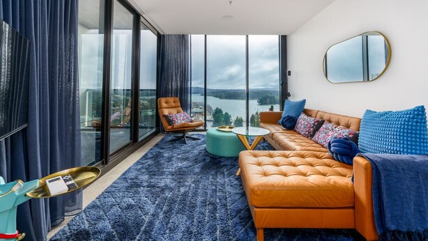 A modern living room with a sofa, armchair, and ottoman, large windows with a view of a body of water, and a round mirror on the wall.