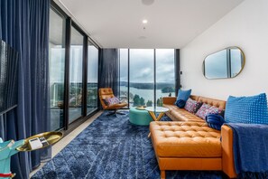 A modern living room with a sofa, armchair, and ottoman, large windows with a view of a body of water, and a round mirror on the wall.
