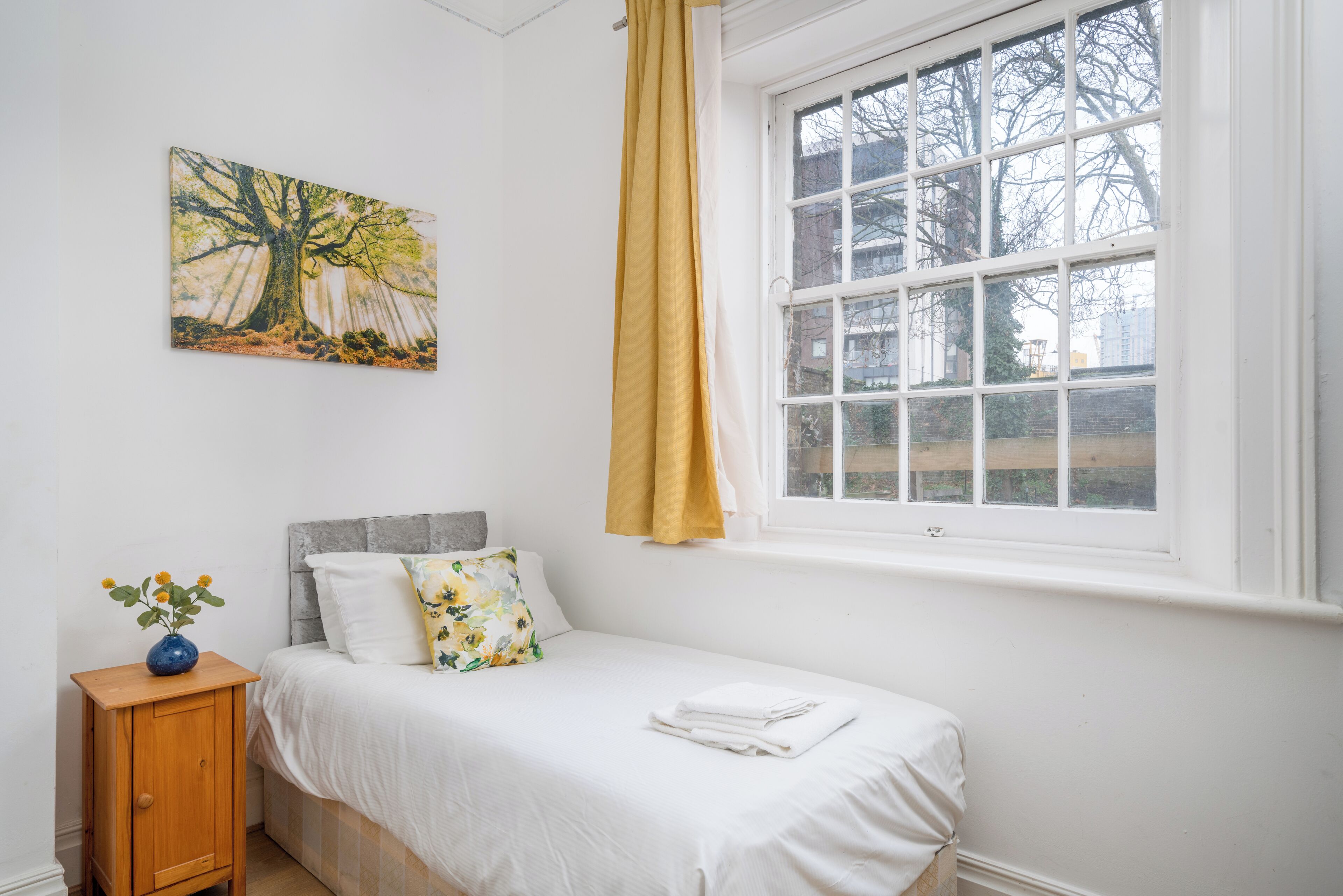 Een slaapkamer met een bed, een nachtkastje met een vaas, een raam met gordijnen en een schilderij aan de muur.