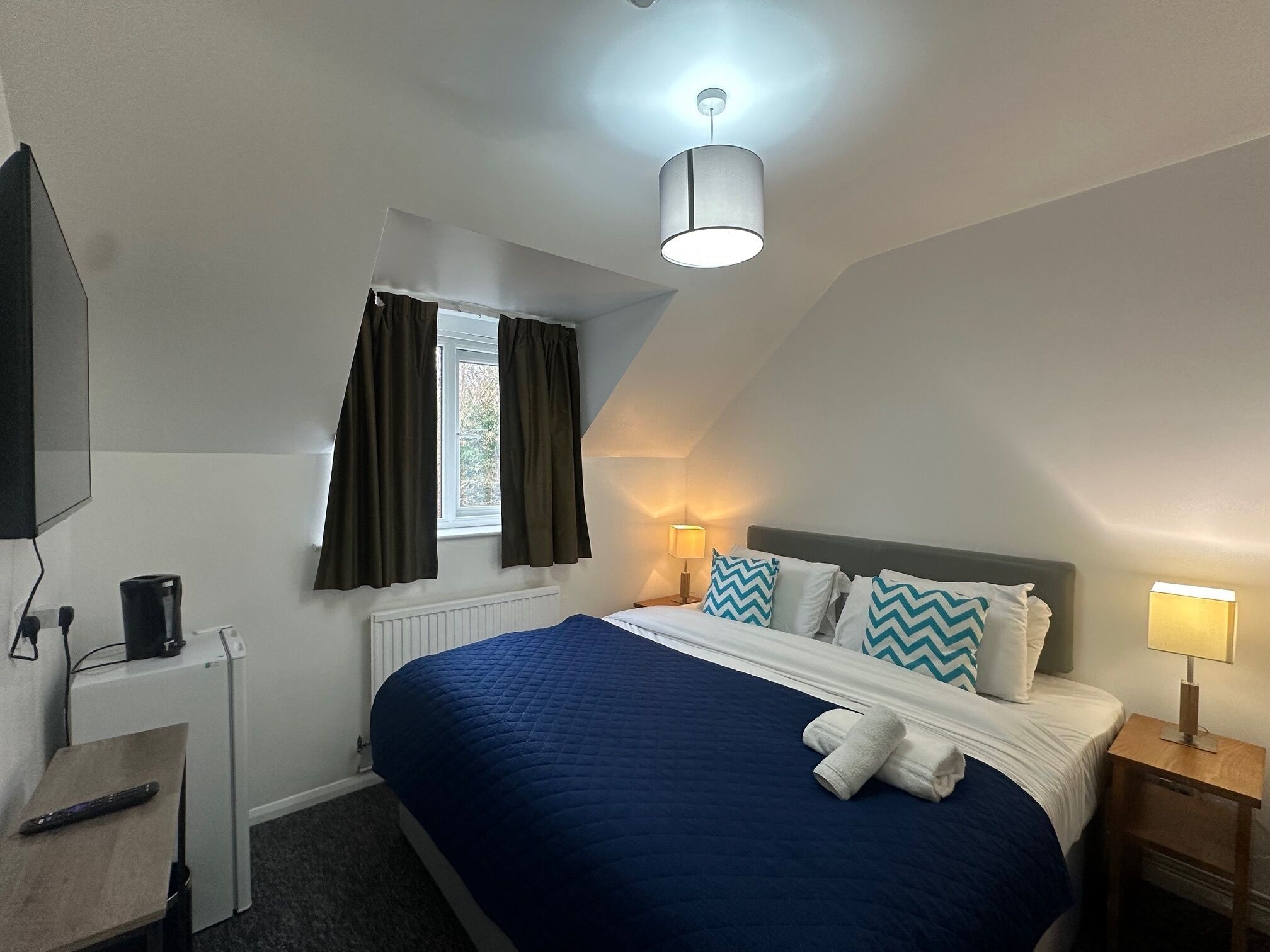 Une chambre d’hôtel comprenant un lit, un bureau, une télévision, une fenêtre avec des rideaux, un luminaire au plafond et une lampe de chevet.