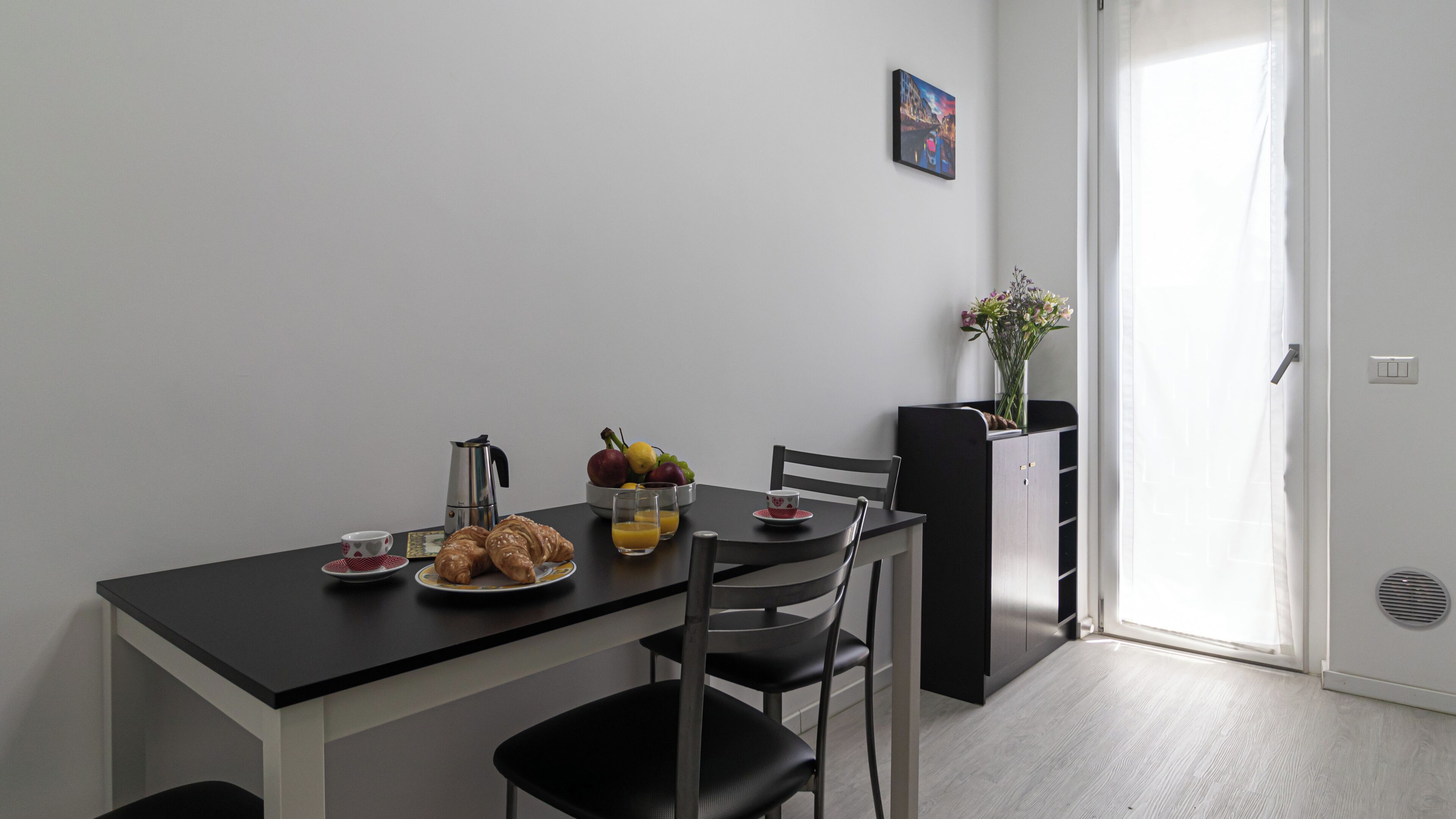 A dining area with a table set for breakfast, including croissants, fruit, and juice. There is a coffee pot, cups, and a vase with flowers on a cabinet.