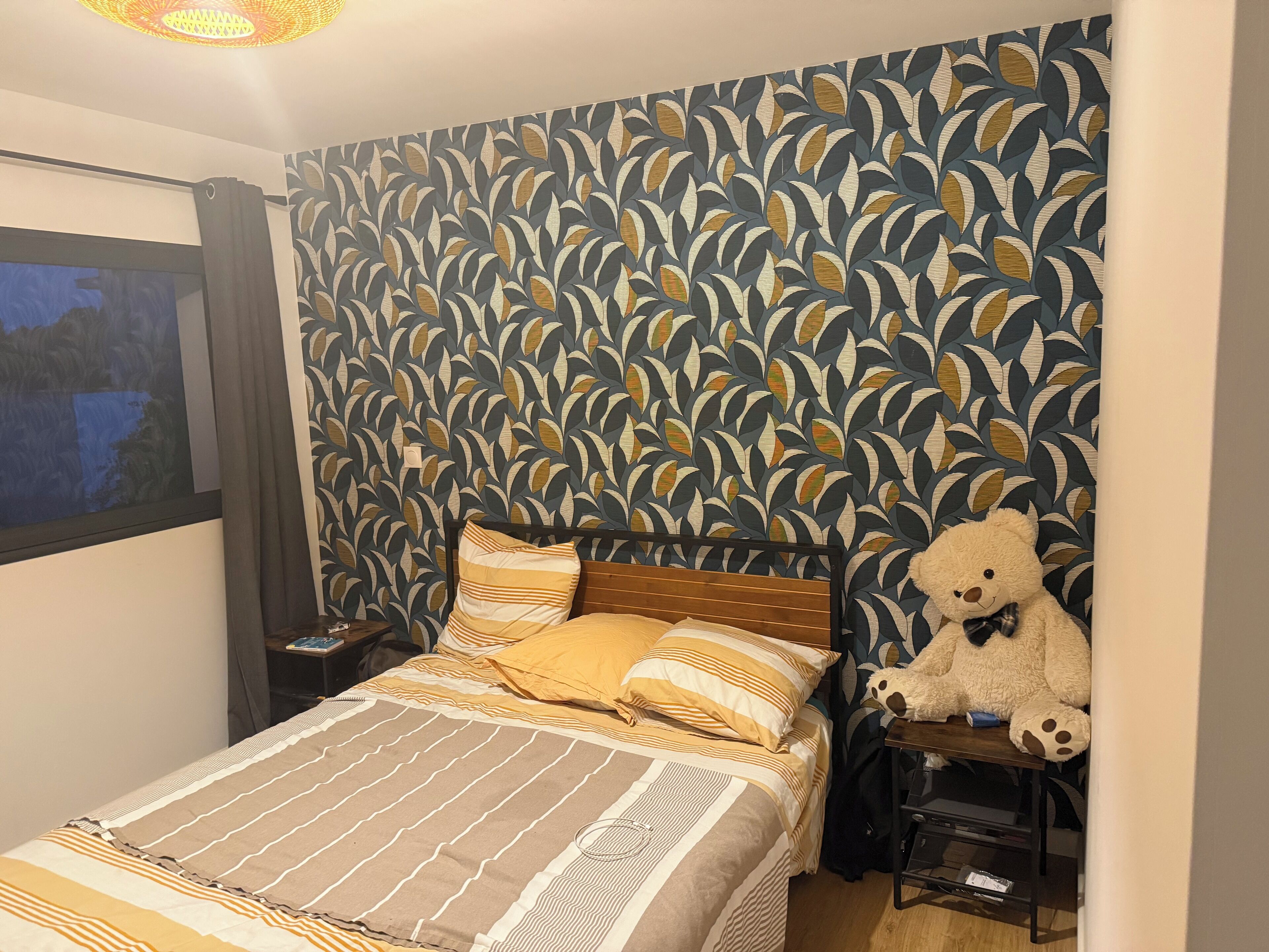 A bedroom with a patterned wallpaper, a bed with striped bedding, and a teddy bear on a nightstand.