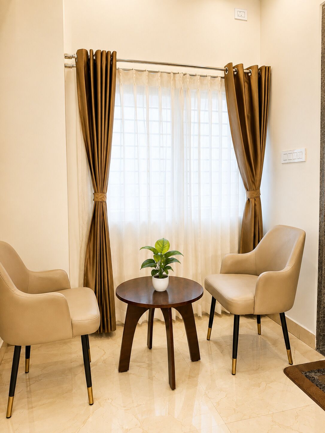 A room with two beige chairs, a small round table with a potted plant, and a window with curtains.