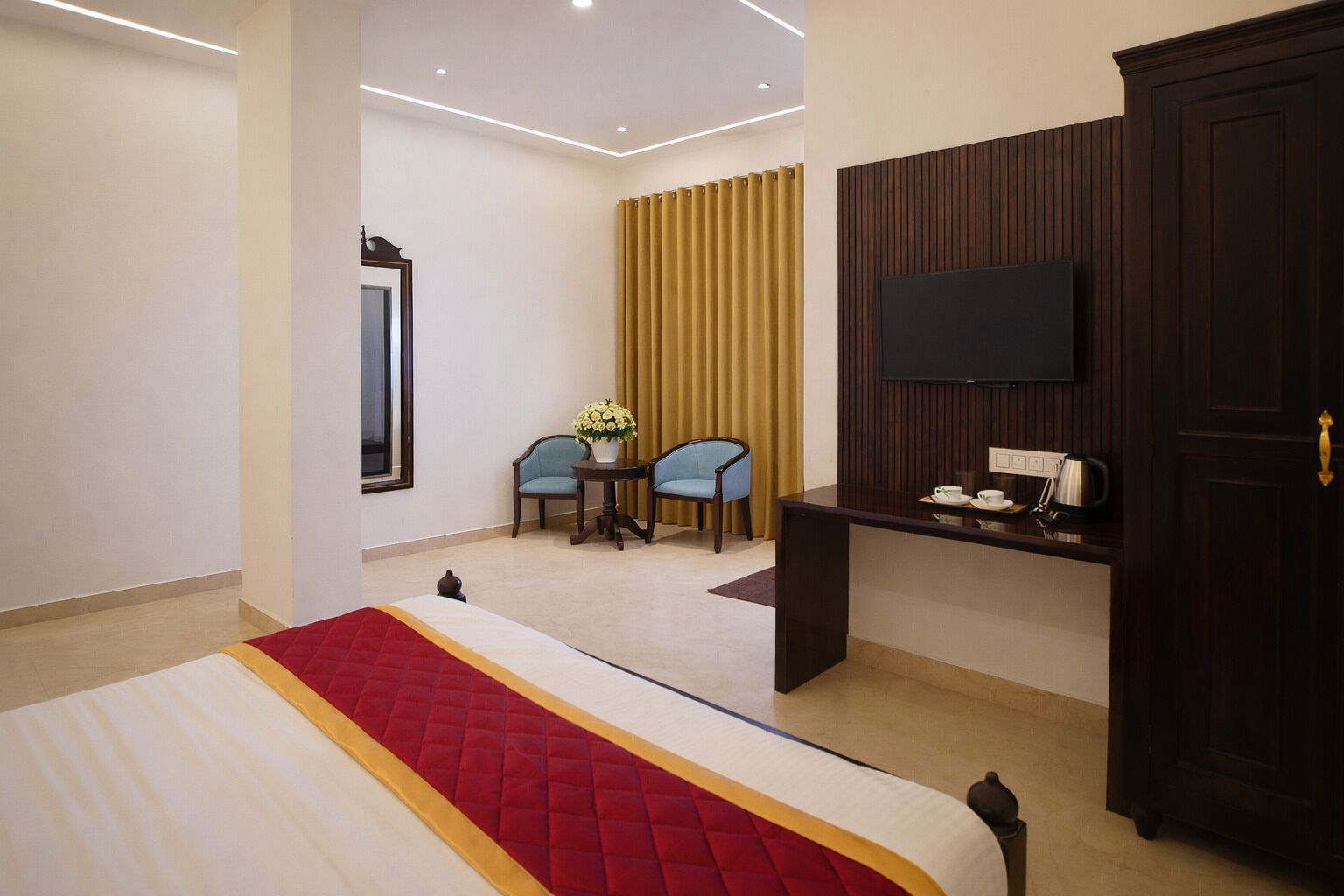 Une chambre d’hôtel comprenant un lit, un bureau avec un téléviseur à écran plat, deux chaises, une petite table avec un vase de fleurs et une armoire.