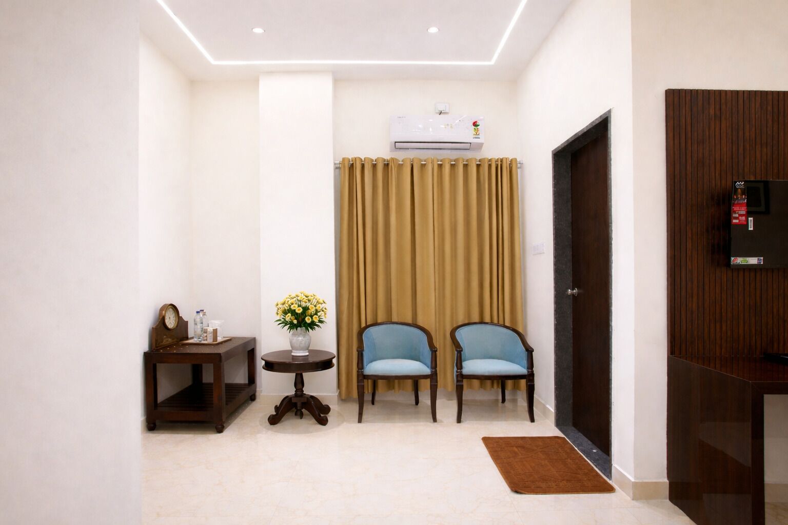 Une chambre d’hôtel avec un lit, un bureau avec une horloge, un vase avec des fleurs, deux chaises, un tapis marron et un appareil de climatisation.