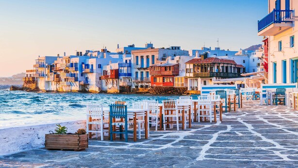 A coastal town with buildings along the waterfront, stone-paved walkways, and outdoor seating areas.