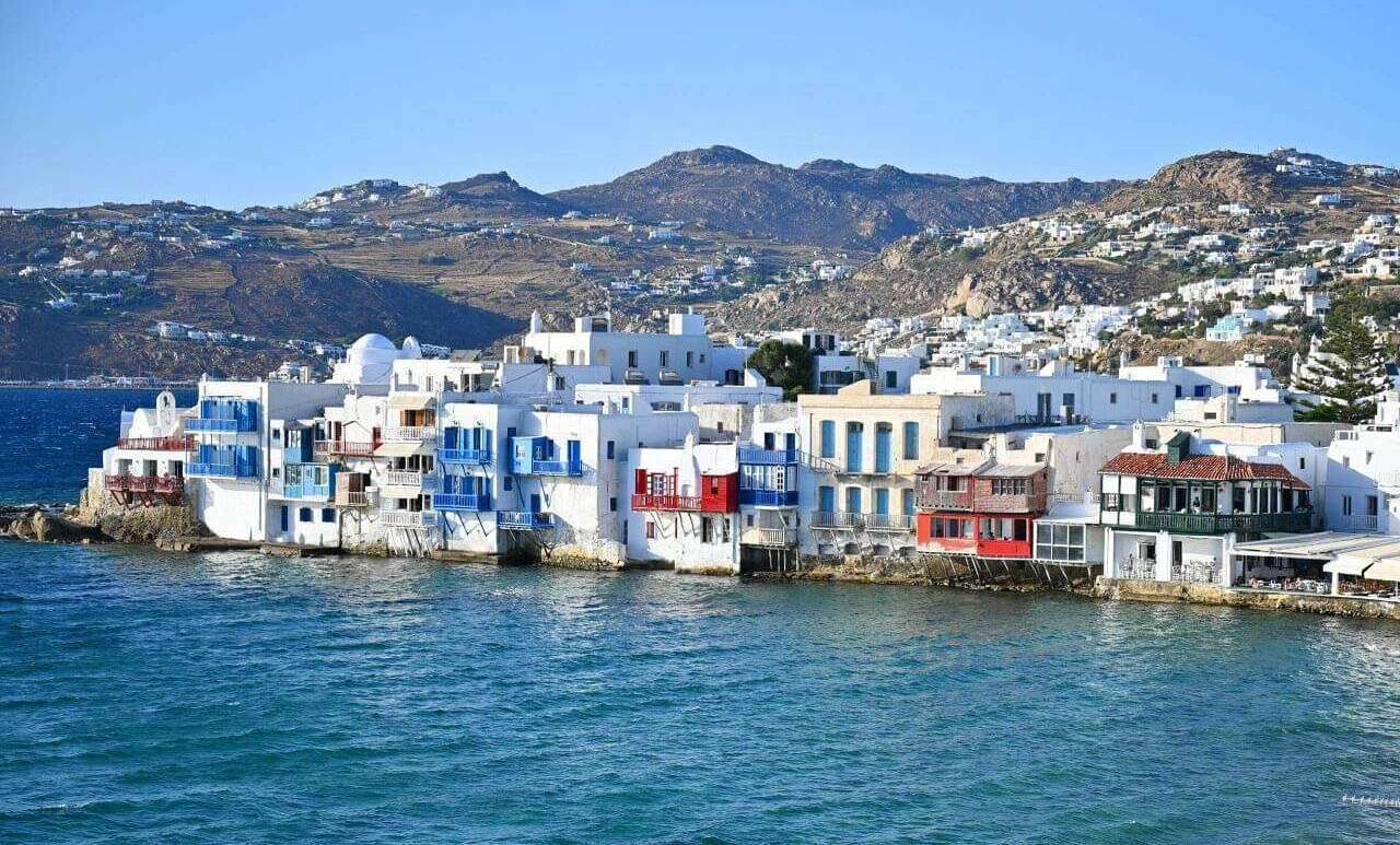 A coastal village with white buildings with blue accents, situated by the sea with a mountainous backdrop.