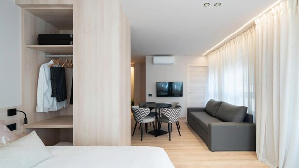 Una habitación de hotel moderna con una cama, una zona de estar con sofá y sillas, un televisor montado en la pared y un armario con ropa colgada.