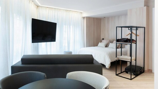 Una habitación de hotel moderna con televisión de pantalla plana, una cama con ropa de cama blanca, una zona de estar con sofá y sillones, y un perchero para equipaje negro.
