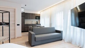 Un salón moderno con un sofá gris, un televisor montado en la pared y una cocina al fondo.