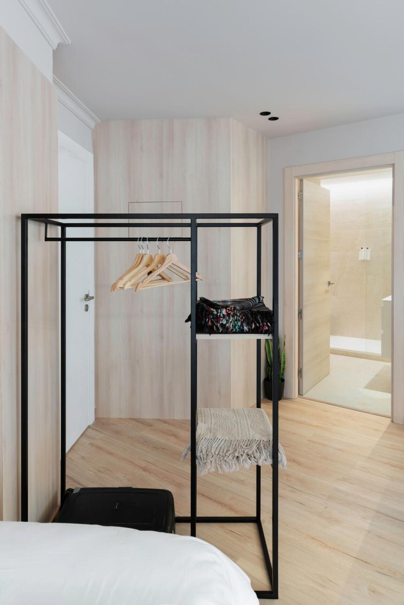 A room with a bed, a black metal wardrobe, wooden hangers, a patterned bag, a small plant, and a suitcase.