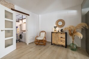 A room with a washing machine, a wicker chair, a wooden cabinet, and a vase with pampas grass.