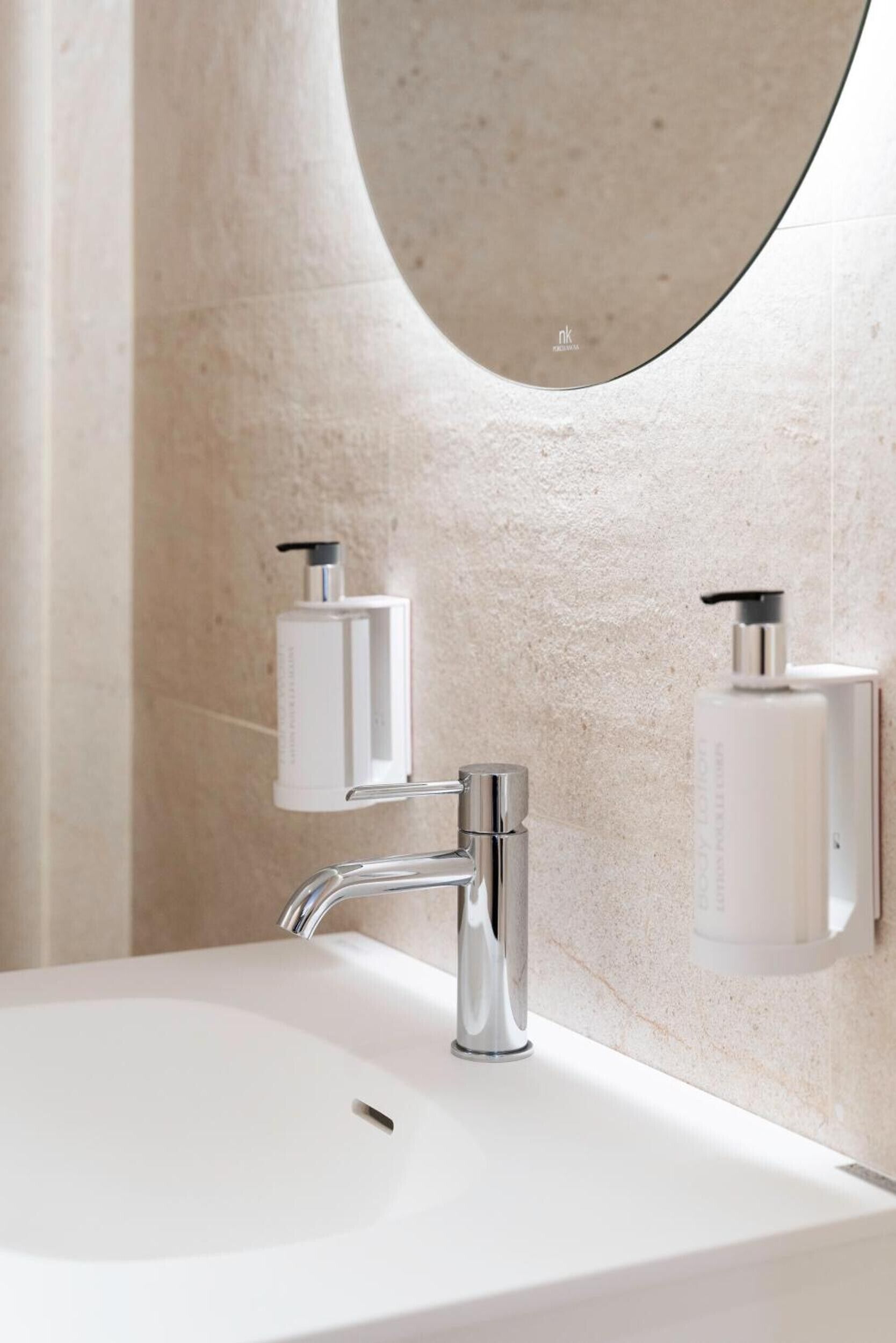Un baño con lavamanos blanco, grifo cromado y un espejo grande. Hay dos dispensadores de jabón blancos montados en la pared.
