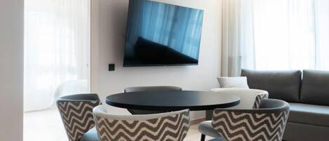 Una sala de estar moderna con una mesa redonda de comedor, cuatro sillas, un televisor de pantalla plana montado en la pared y un sofá gris.