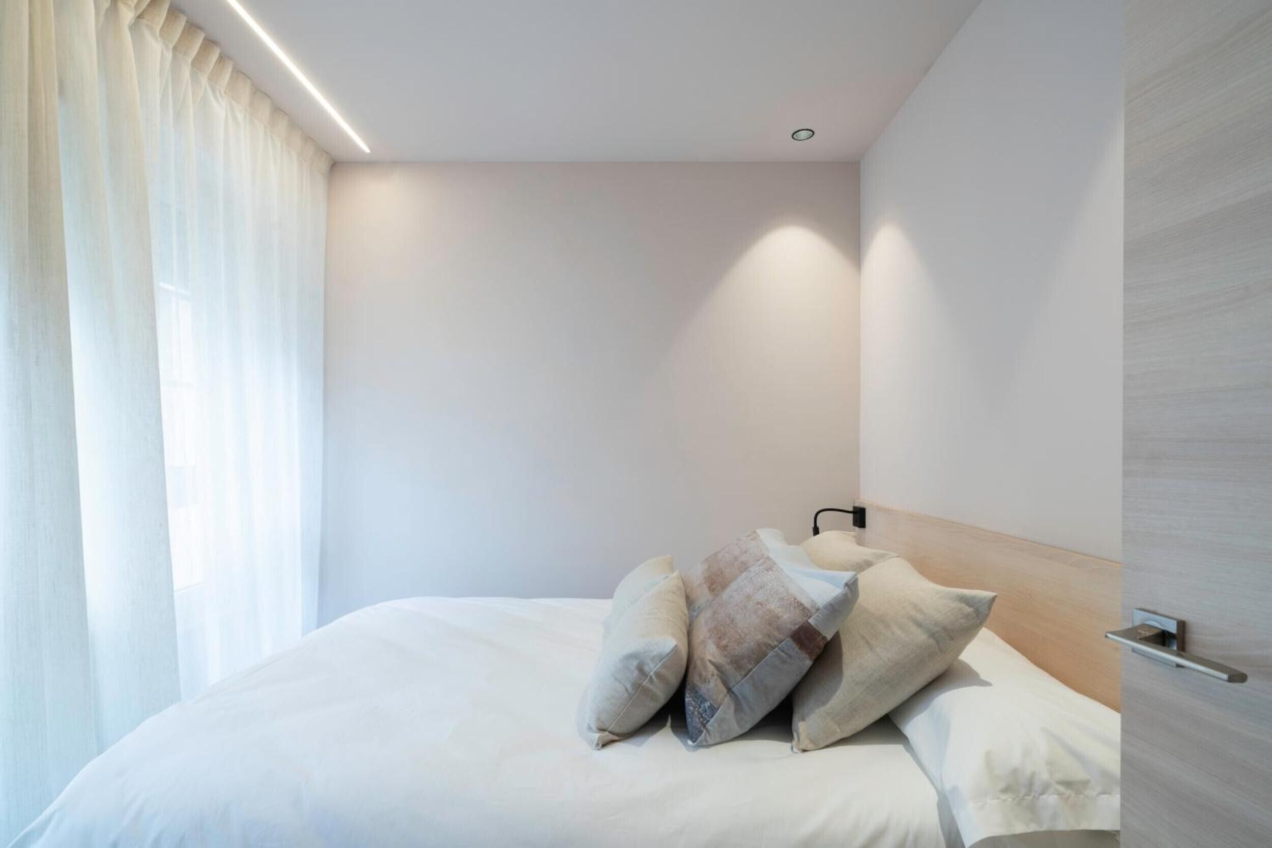 Moderni hotellihuone, jossa on siististi pedattu sänky, puinen pääty ja ikkuna, jossa on ohuet verhot.