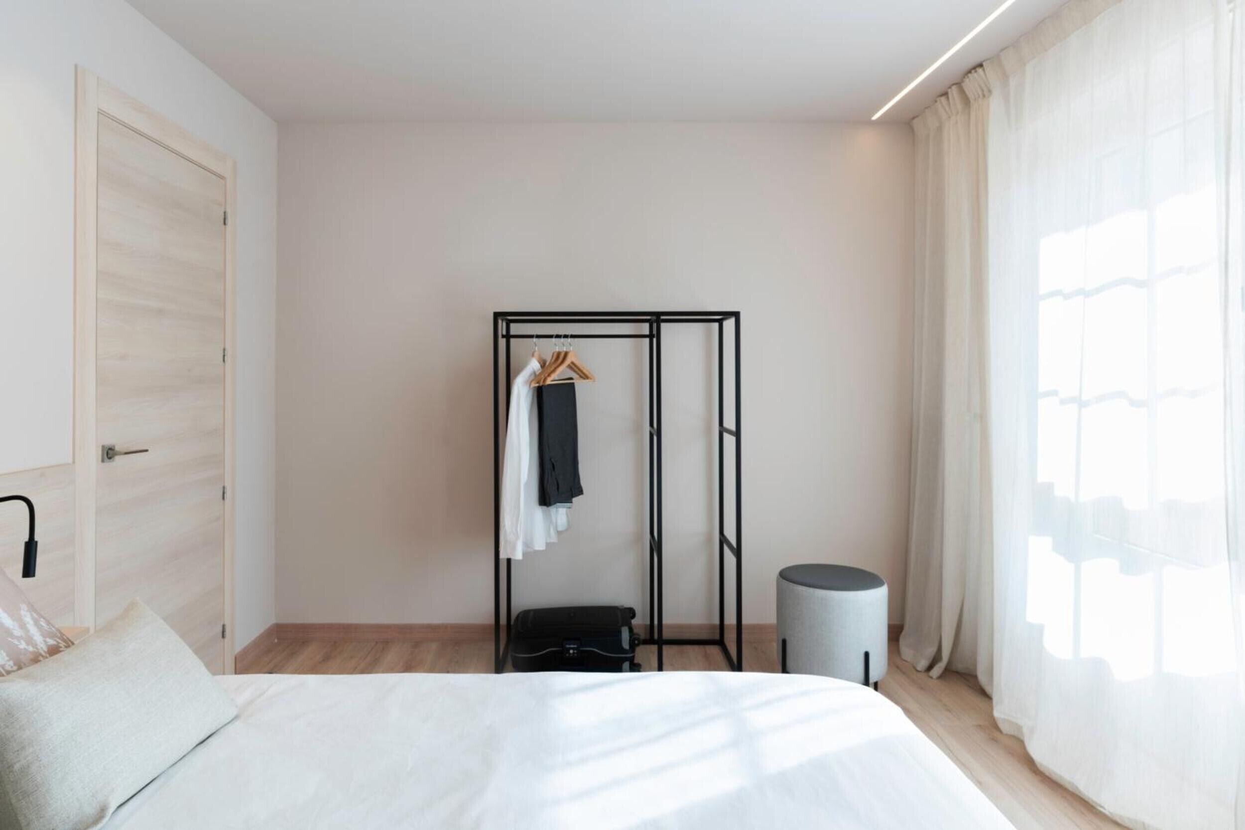 Un dormitorio con cama, armario, perchero, cubo de basura y ventana con cortinas.