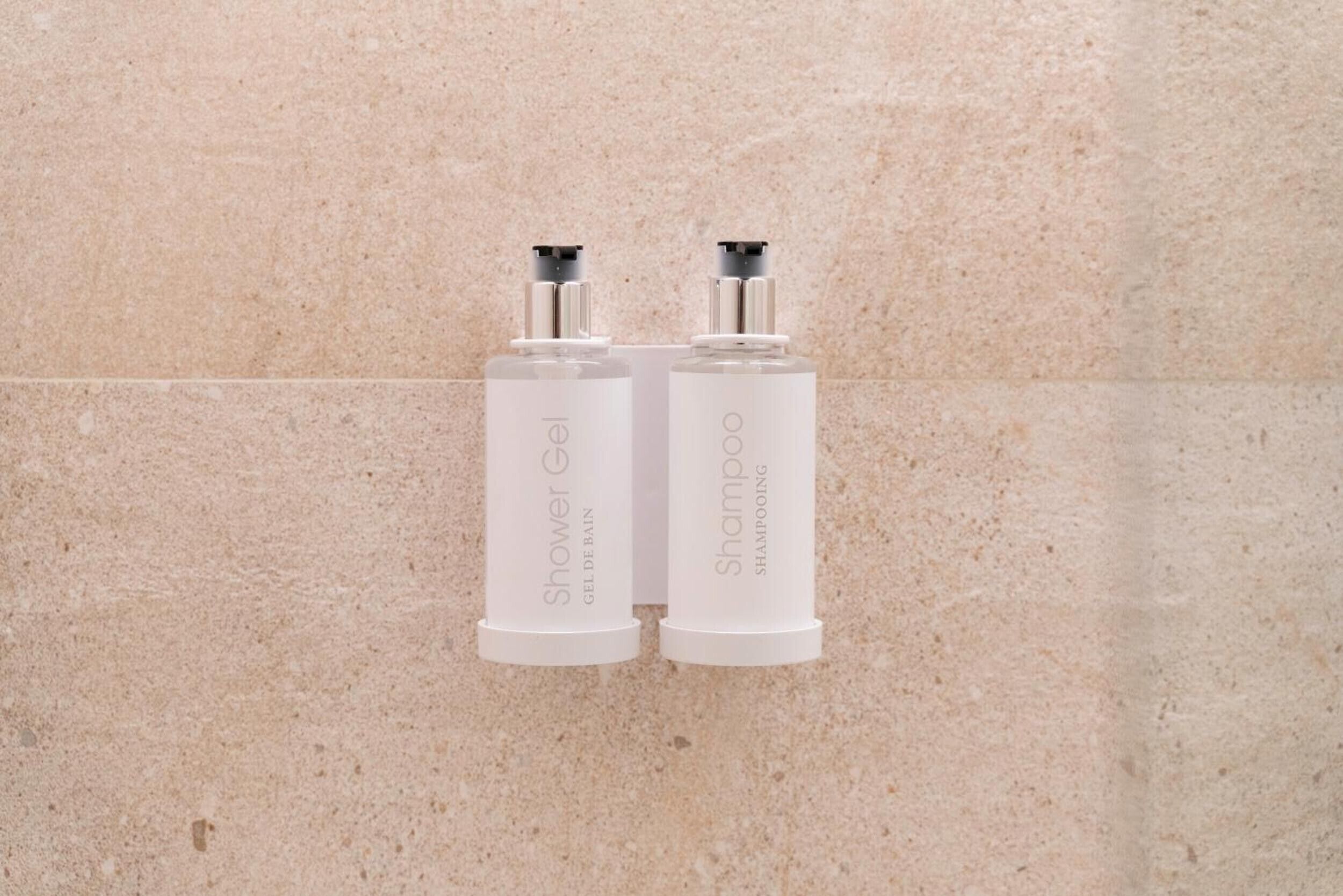 Dos botellas blancas etiquetadas 'Shower Gel' y 'Shampoo' sujetas a una pared de azulejos color beige.