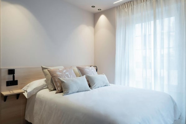 A neatly made bed with white bedding and multiple pillows, a bedside table with a lamp, and sheer curtains on a window.