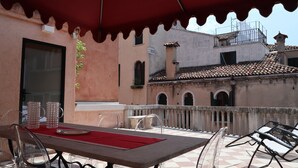 Patio esterno con tavolo apparecchiato per mangiare, un ombrello rosso e vista su un edificio con tetto in tegole.