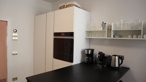 Un angolo cottura con un piano di lavoro nero, una macchina da caffè, un bollitore e un forno a microonde. Sono presenti mobili bianchi e uno scaffale con bicchieri.