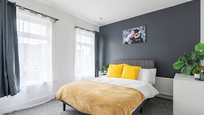 Un dormitorio moderno con cama, cabecera gris, cuadro abstracto colorido, planta en maceta y radiador.