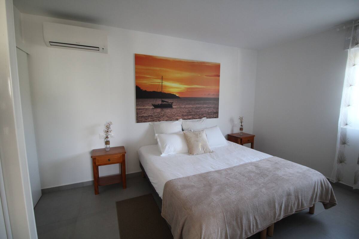 Een slaapkamer met een bed, twee nachtkastjes, een schilderij van een ondergaande zon boven water, en een raam met gordijnen.