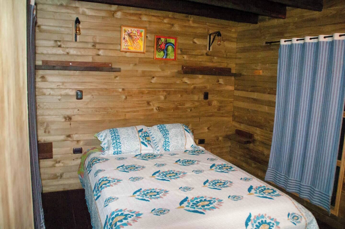 Una habitación de cabaña de madera con una cama, dos lámparas de pared y dos obras de arte enmarcadas.
