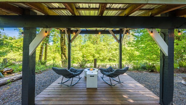 A wooden deck with two rocking chairs and a fire pit, surrounded by a covered pergola and lush greenery. - Private Riverfront home w/ Hot tub, central to ONP (Beaver)