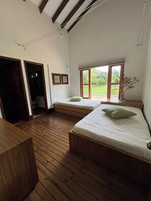 Une chambre avec deux lits, un plancher en bois, une grande fenêtre et une porte donnant sur une autre pièce.