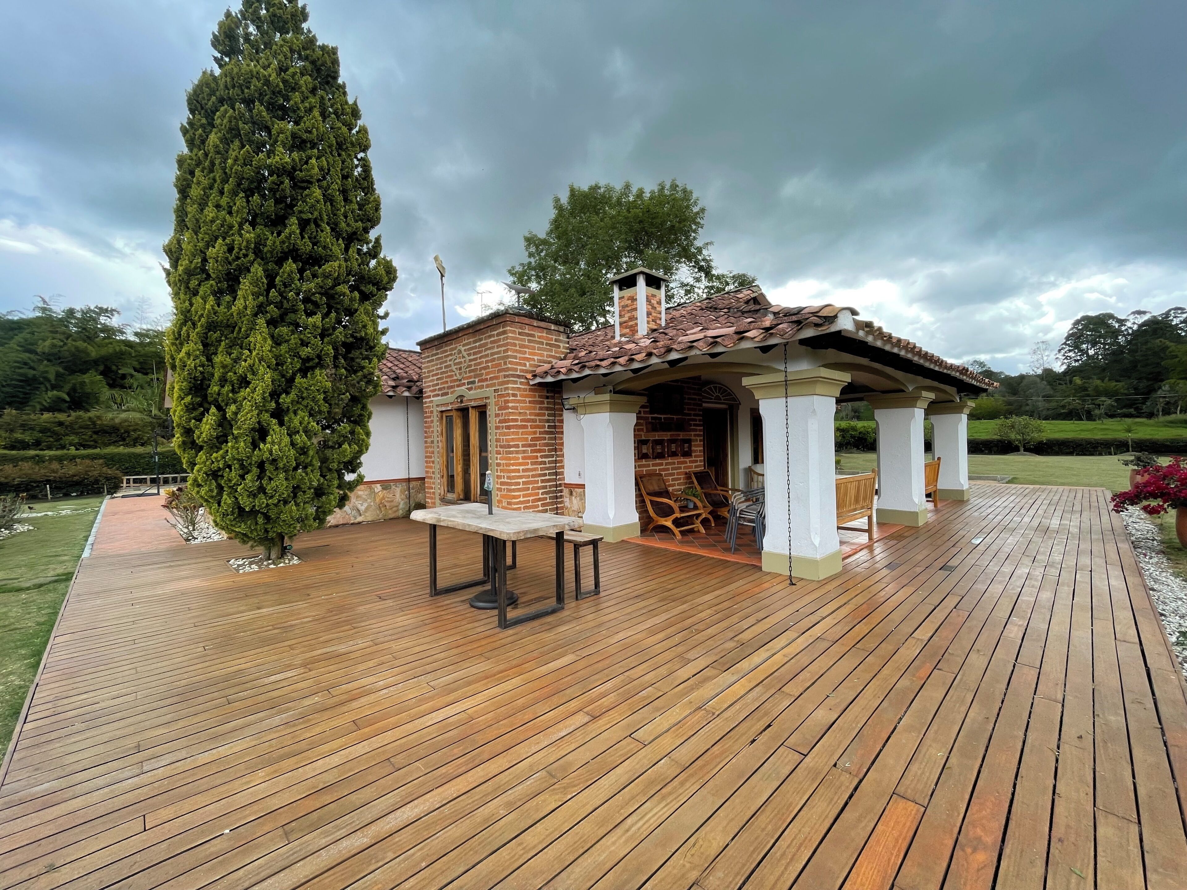 Une terrasse en bois avec un espace patio couvert, un bâtiment en briques et un grand arbre à feuilles persistantes.