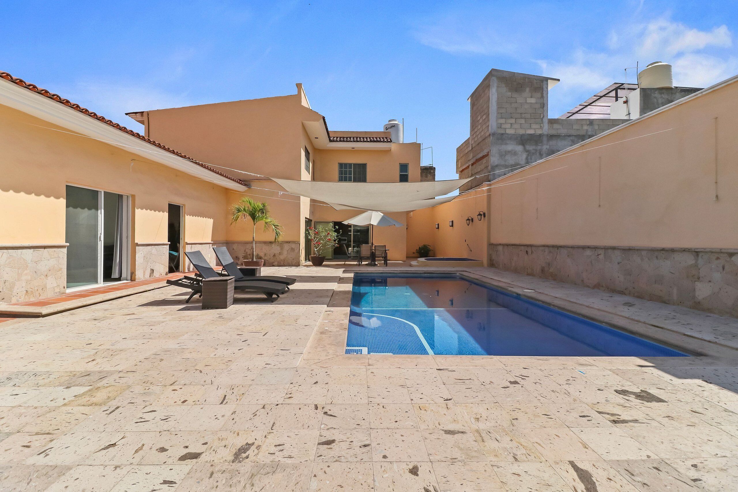 Una piscina con sillas de descanso y una zona con sombra, rodeada de edificios.