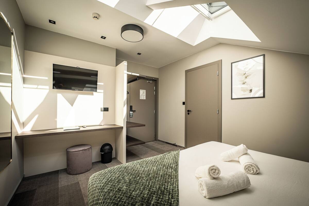 Une chambre d’hôtel moderne avec une lucarne, un lit avec des serviettes, une poubelle et une porte donnant sur une autre chambre.
