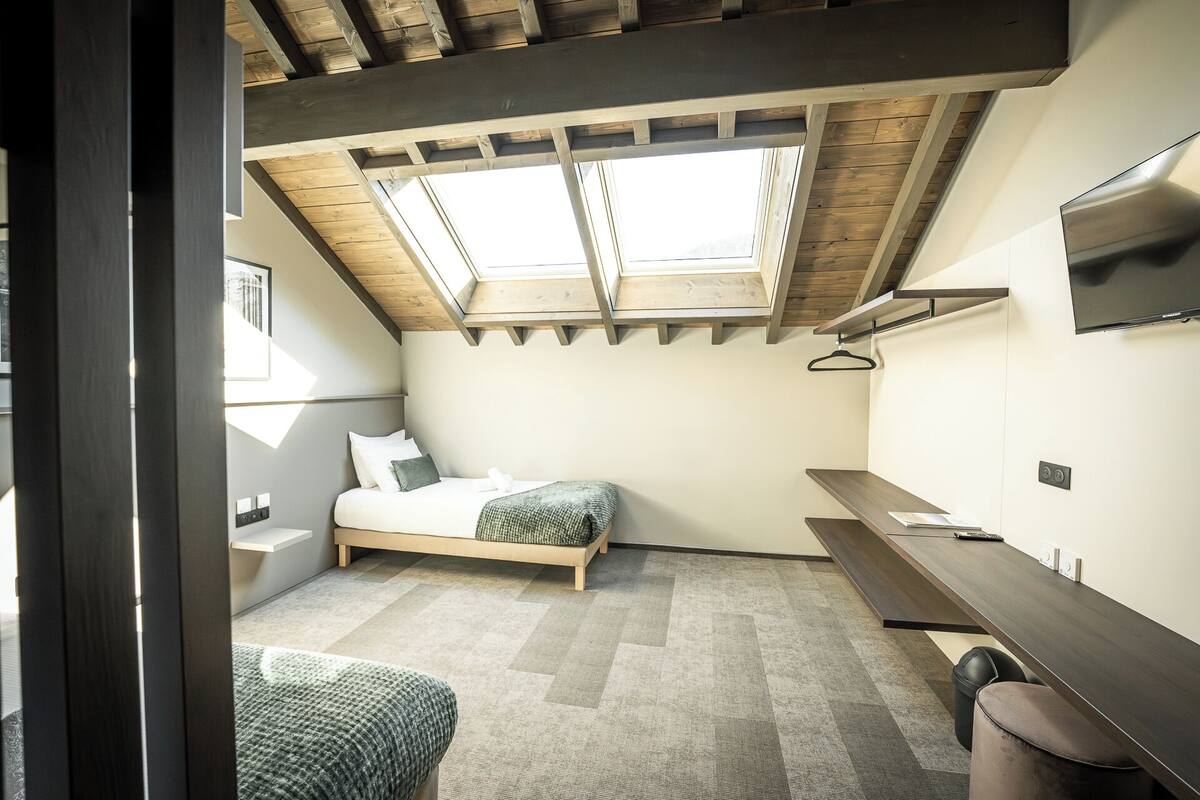 Une petite pièce comprenant un lit, un bureau, une télévision et une lucarne.