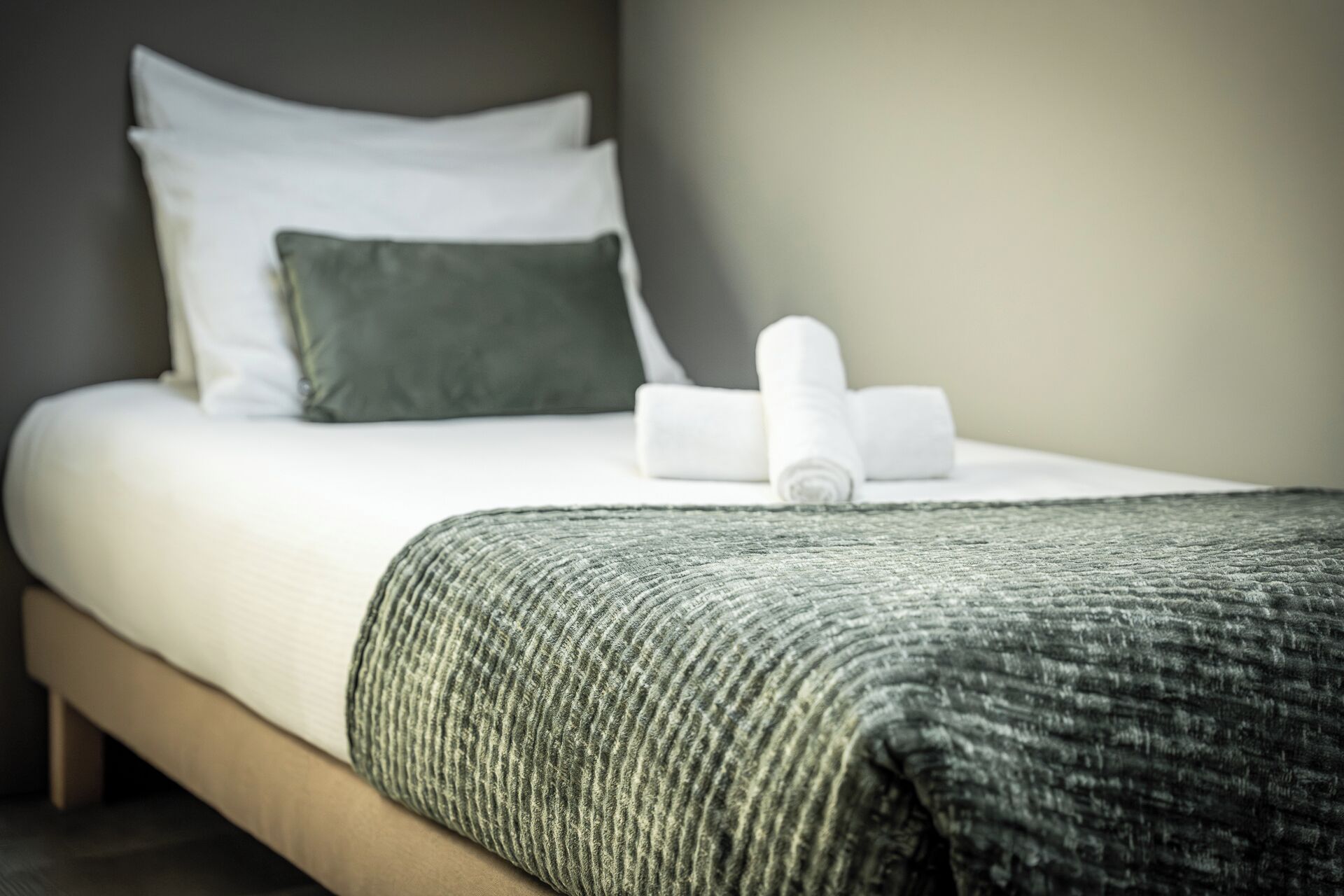 A neatly made bed with white linens, a green pillow, and a rolled white towel.
