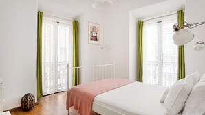A bedroom with a bed, a bedside lamp, a window with green curtains, and a door with a white frame.