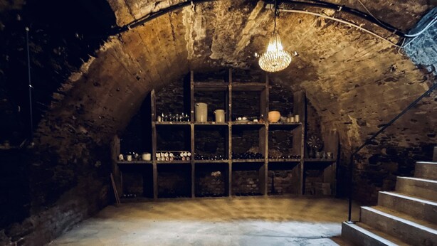 A dimly lit stone cellar with a chandelier, wooden shelves, and a staircase. - Former wine château (Winningen)