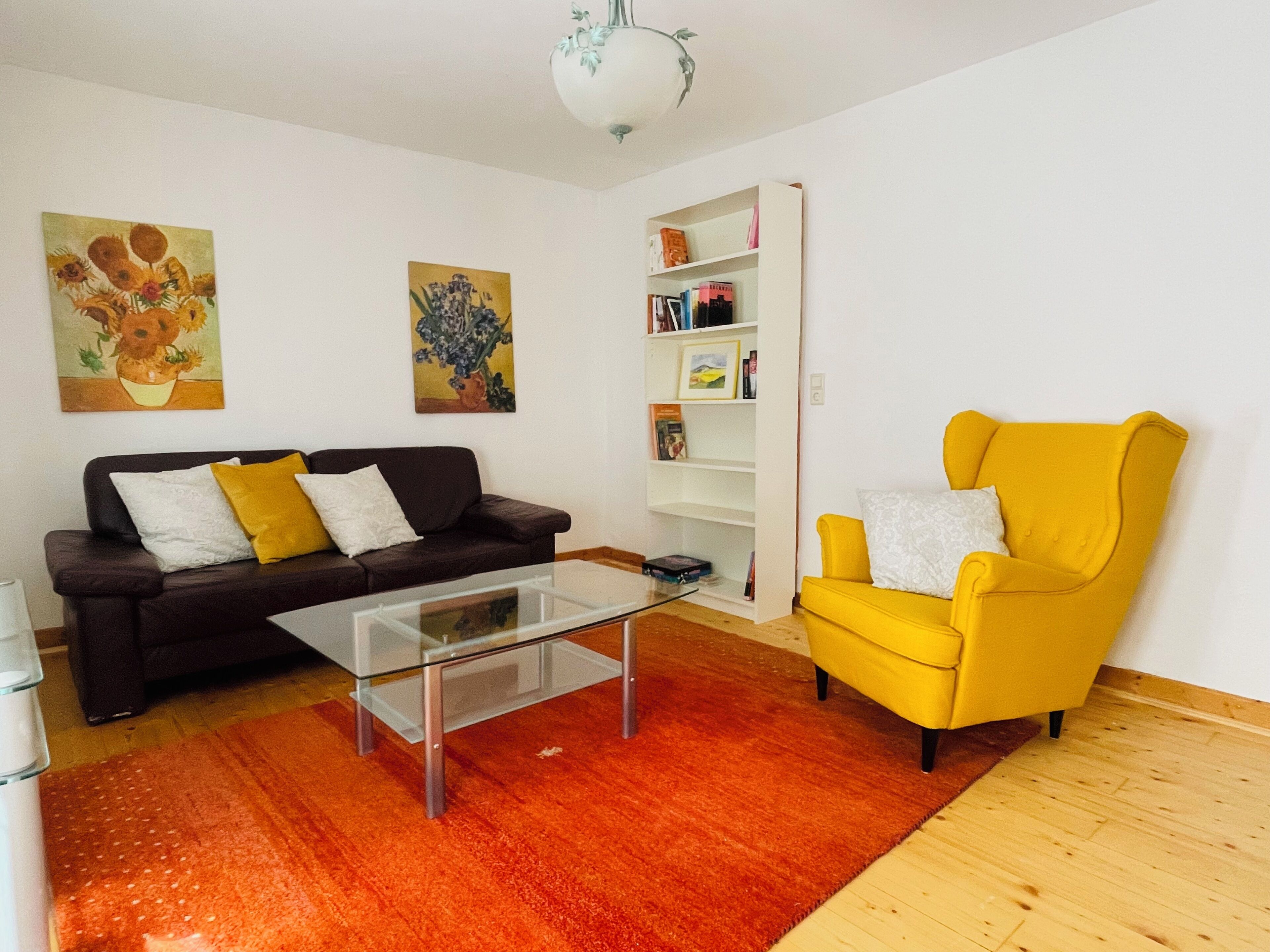 Una sala de estar con un sofá marrón, un sillón amarillo, una mesa de centro de vidrio y una estantería.