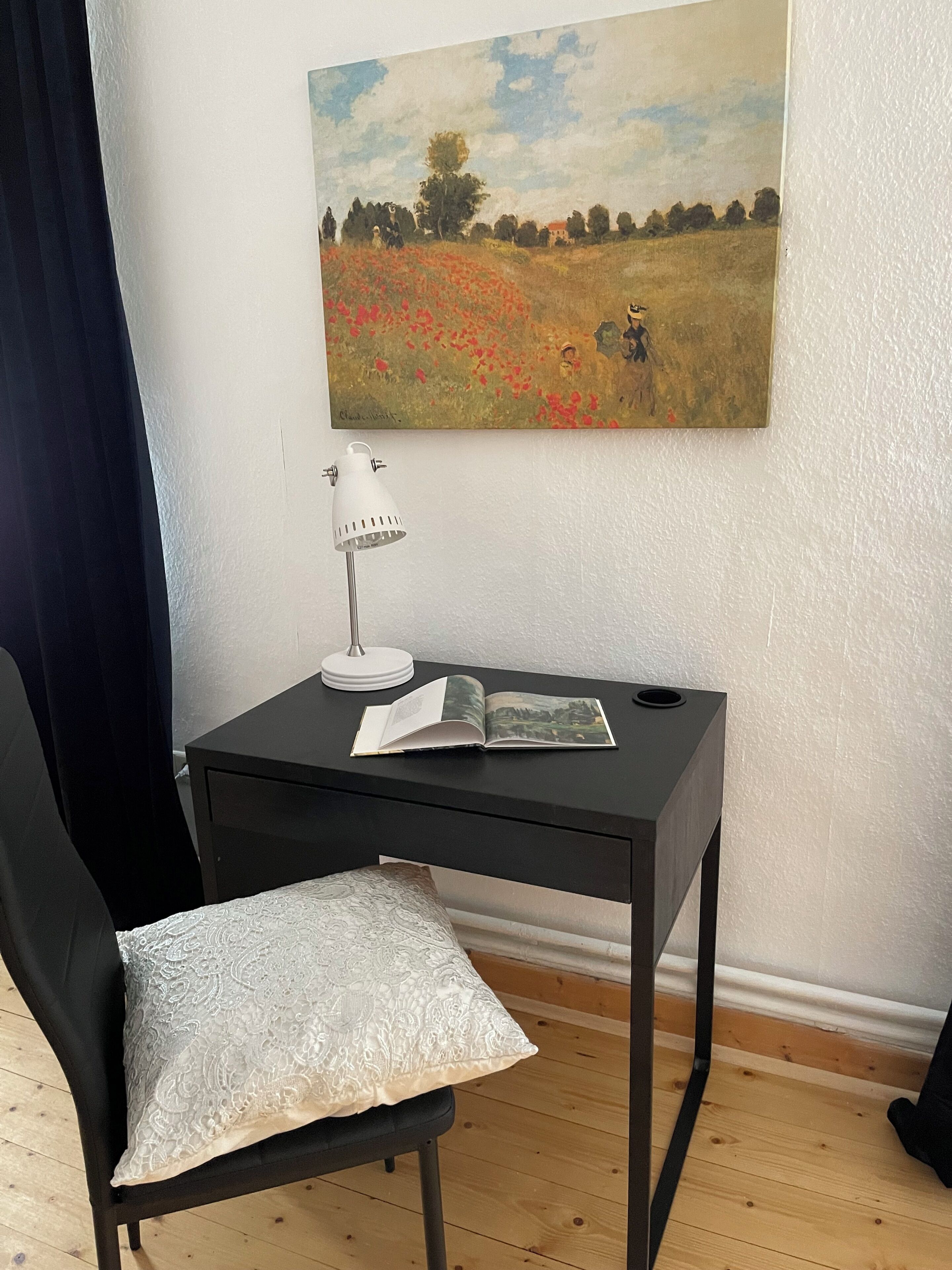 A room with a black desk, a chair with a patterned cushion, a lamp, and a book. A painting of a field with red flowers hangs on the wall.