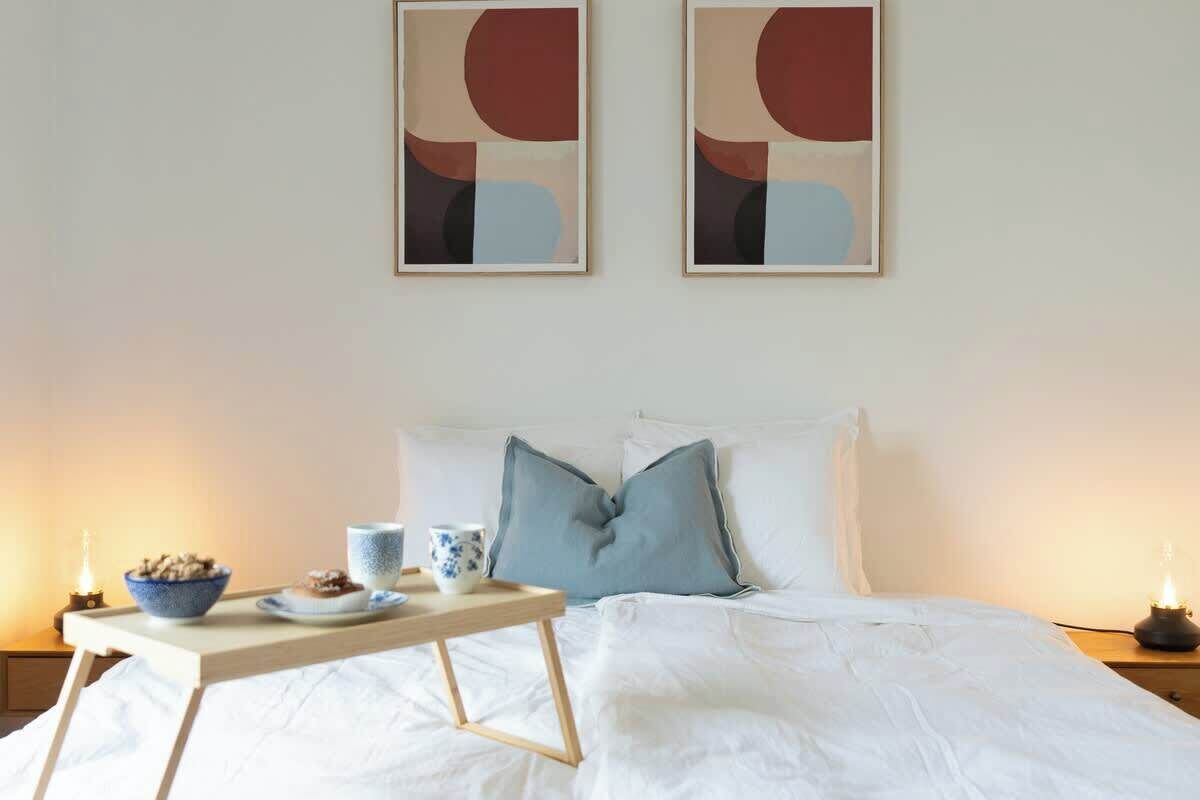 A neatly made bed with pillows, a bedside table with a bowl of cereal and a cup, and two abstract paintings on the wall.