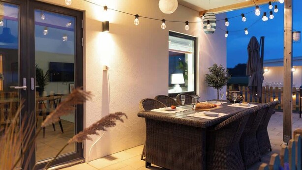 Un espacio de comedor al aire libre con mobiliario de mimbre, luces colgantes y un ventanal que da al interior.