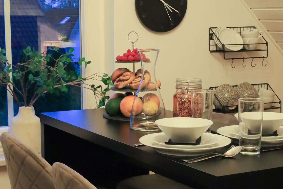 Zona de menjador amb taula negra preparada per a quatre, incloent bols, plats i coberts. Hi ha envasos de vidre amb aliments i una presentació de fruita sobre la taula.
